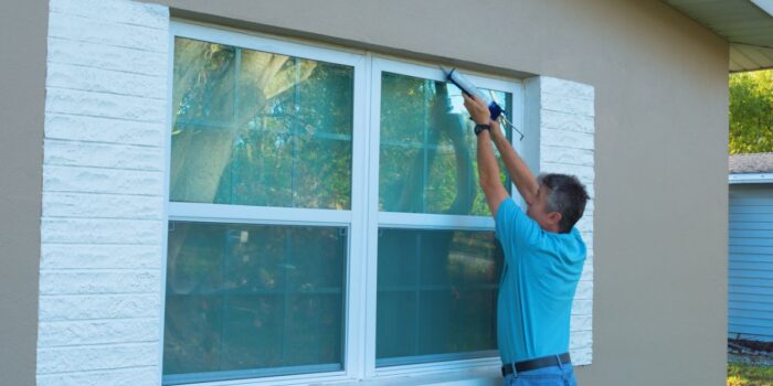 A man in a blue shirt is sealing storm windows on a house, an activity that raises the question "what are storm windows" in relation to their role in protecting homes from severe weather.